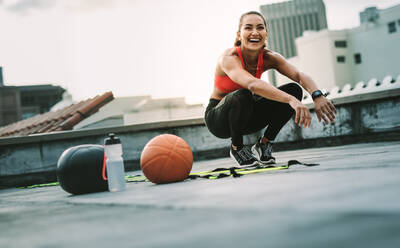 Fröhliche Frau in Fitnesskleidung beim Workout auf der Terrasse. Fitness-Frau macht Sit-ups auf dem Dach mit Medizinball und Basketball an ihrer Seite. - JLPSF19715