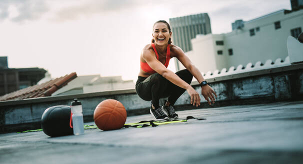 Fitness-Frau sitzt auf ihren Zehen, während sie auf dem Dach trainiert. Fröhliche Frau Athleten tun Training auf dem Dach mit Medizinball und Basketball an ihrer Seite. - JLPSF19716