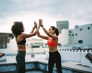 Zwei Fitness-Frauen feiern, indem sie nach dem Training High Five geben. Fitness-Frauen, die ihr Training auf dem Dach genießen und High Five mit beiden Händen geben. - JLPSF19720