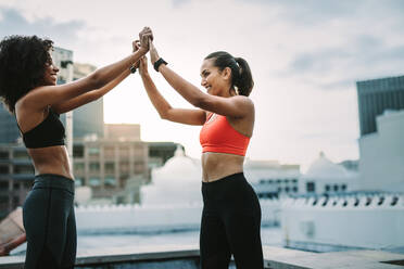 Happy Fitness Frauen geben High Five nach ihrem Training auf dem Dach. Zwei Frauen in Fitness-Kleidung stehen auf dem Dach in freudiger Stimmung nach dem Training. - JLPSF19721