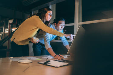 Eine Geschäftsfrau zeigt ihrem Geschäftspartner etwas auf dem Computerbildschirm. Ein Geschäftsmann und eine Frau arbeiten spät in der Nacht im Büro zusammen. - JLPSF19724