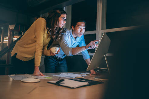 Ein Geschäftsmann und eine Frau besprechen ihre Arbeit, während sie spät in der Nacht auf einen Computer schauen. Geschäftskollegen arbeiten ernsthaft spät in der Nacht im Büro, um Arbeitsfristen einzuhalten. - JLPSF19725