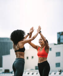 Seitenansicht von zwei Fitness-Frauen, die auf dem Dach stehend High Five geben. Fitness-Frauen, die ihr Training auf dem Dach genießen und High Five mit beiden Händen geben. - JLPSF19727
