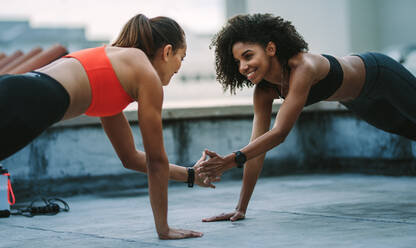 Lächelnde Fitness-Frauen machen Liegestütze auf einem Dach und stehen sich gegenüber. Frauen berühren ihre Hand, während sie gleichzeitig mit der anderen Hand Liegestütze machen. - JLPSF19730