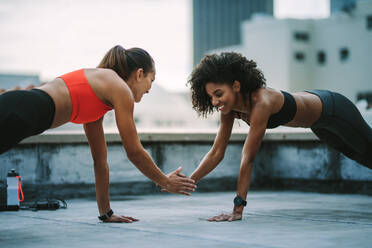 Zwei Fitness-Frauen machen Liegestütze mit einer Hand, während sie ihre andere Hand berühren. Fröhliche Frauen machen Fitness-Workout zusammen auf dem Dach. - JLPSF19731