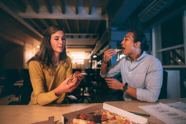 Geschäftskollegen, die spät in der Nacht im Büro zusammensitzen und Pizza essen. Mann und Frau, die spät im Büro sitzen und nach getaner Arbeit etwas essen. - JLPSF19732