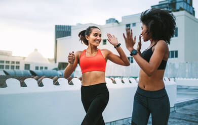 Zwei Fitness-Frauen joggen auf dem Dach am Morgen und geben high five. Frauen Athleten in fröhlicher Stimmung tun Fitness-Training auf dem Dach. - JLPSF19737