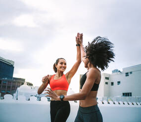 Zwei fröhliche Frauen in Fitnesskleidung geben sich beim Laufen auf der Terrasse ein High Five. Sportlerinnen beim Fitnesstraining auf dem Dach geben sich ein High Five. - JLPSF19738