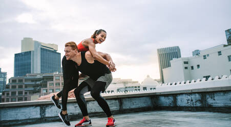 Mann hebt eine Sportlerin auf seinen Rücken während des Fitnesstrainings auf dem Dach. Fitness-Mann und Frau in fröhlicher Stimmung haben Spaß beim Training auf der Terrasse. - JLPSF19739