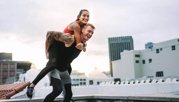 Athlete carrying a fitness woman on his back and walking on rooftop. Smiling fitness woman piggy riding on a man while training on rooftop. - JLPSF19740