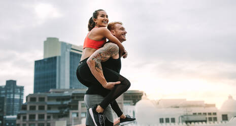 Fitness-Paar, das sich beim Training auf dem Dach amüsiert. Lächelnde Sportlerin, die beim Training auf dem Dach huckepack auf einem Mann reitet. - JLPSF19741