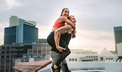 Seitenansicht eines Fitness-Paares, das sich beim Training auf einer Gebäudeterrasse amüsiert. Ein Mann, der eine Fitness-Frau auf seinem Rücken trägt, läuft auf einem Dach. - JLPSF19742