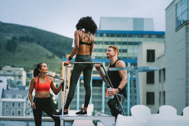 Fitness-Mann steht mit weiblichen Athleten, die eine Wasserflasche halten und sich auf einem Dach unterhalten. Mann und Frauen in Fitnesskleidung machen eine Pause nach dem Training auf einer Terrasse. - JLPSF19743