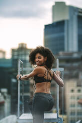 Afroamerikanische Frau in Fitnesskleidung klettert die Treppe auf dem Dach hinunter. Fitnessfrau steht auf dem Dach und schaut zurück. - JLPSF19749