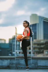 Smiling fitness woman standing near a rooftop staircase with a ball in hand. Rear view of a woman in fitness wear standing on rooftop doing fitness training. - JLPSF19752