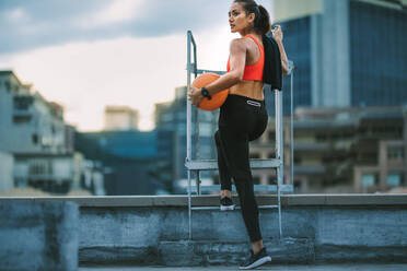 Woman in fitness wear standing on rooftop holding a basketball. Rear view of a fitness woman standing near a rooftop staircase and looking away. - JLPSF19753