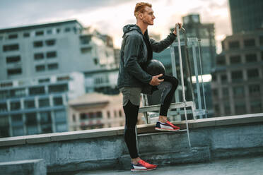 Sportler, der mit einem Medizinball in der Hand auf einer Dachtreppe steht. Seitenansicht eines Mannes in Fitnesskleidung, der auf einem Dach Fitnessübungen macht. - JLPSF19754