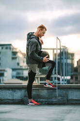 Fitness man standing on rooftop holding a medicine ball. Side view of a relaxed athlete standing near a rooftop staircase and looking down. - JLPSF19755