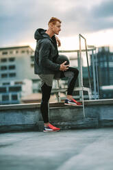 Side view of an athlete standing near a rooftop staircase and looking away. Man in fitness clothes standing relaxed holding a medicine ball on rooftop stairs. - JLPSF19756