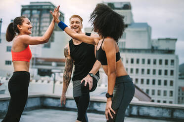 Zwei Fitness-Frauen drücken ihre Freude aus, indem sie sich nach dem Training ein High Five geben, während ein Mann zusieht. Fitness-Mann und Frauen entspannen sich und haben Spaß nach dem Training auf dem Dach. - JLPSF19762