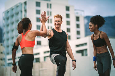 Weiblicher Athlet, der einem Mann nach dem Fitnesstraining auf dem Dach stehend High Five gibt. Fitness-Mann und Frauen in fröhlicher Stimmung geben High Five. - JLPSF19763