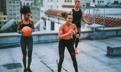 Lächelnde Fitness-Frauen und ein Mann beim Fitnesstraining auf der Terrasse. Ein Mann trainiert mit einem Springseil, während seine Kolleginnen mit einer Agility-Leiter und einem Basketball trainieren. - JLPSF19766