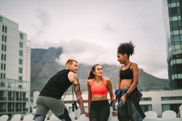 Zwei Fitness-Frauen mit einem Mann entspannen sich nach dem Training auf dem Dach und reden miteinander. Fitness-Leute stehen auf der Terrasse nach dem Fitnesstraining an einem angenehmen bewölkten Morgen. - JLPSF19770