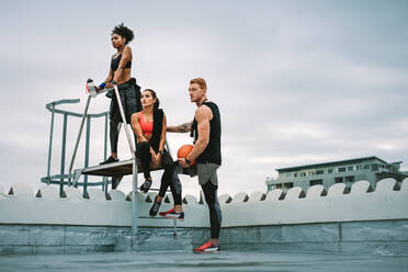 Man holding a basketball standing with two female athletes on rooftop stairs after fitness training. Low angle view of three fitness people relaxing after workout near rooftop stairs and looking away. - JLPSF19773