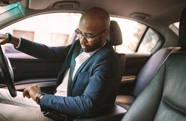Junger afrikanischer Geschäftsmann, der ein Auto fährt und die Zeit überprüft. Männlicher Fahrer sitzt im Auto im Stau und schaut auf seine Armbanduhr. - JLPSF19777