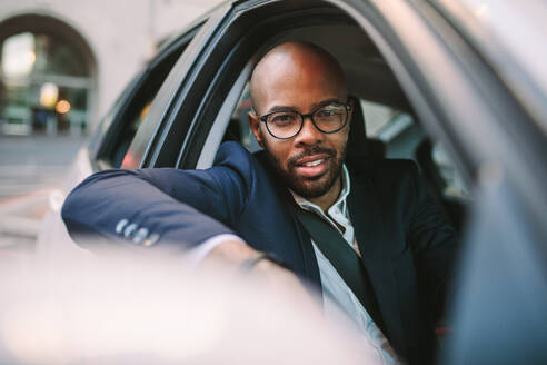 Afrikanischer Geschäftsmann im Anzug, der während der Fahrt aus dem Autofenster schaut und in die Kamera blickt. Gutaussehender Geschäftsmann im Anzug, der auf einer Stadtstraße Auto fährt. - JLPSF19780