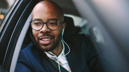 Nahaufnahme eines glücklichen jungen Geschäftsmannes mit Kopfhörern, der während der Fahrt aus dem Autofenster schaut. Afrikanischer Geschäftsmann im Anzug fährt mit dem Auto ins Büro. - JLPSF19783