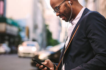 Fröhlicher Millennial-Geschäftsmann beim Pendeln mit einem Smartphone. Afrikanischer Geschäftsmann, der Musik über seine Kopfhörer hört, während er im Freien auf einer Stadtstraße spazieren geht. - JLPSF19787