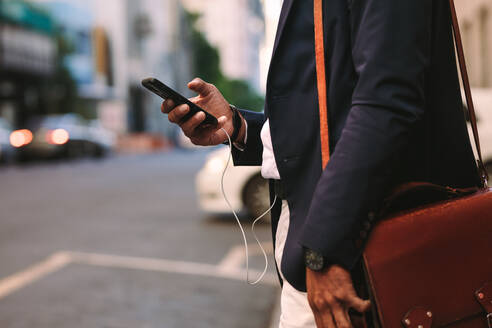 Ausschnitt eines Geschäftsmannes, der im Freien spazieren geht und dabei sein Handy benutzt. Mann im Anzug mit Handtasche, der auf einer Straße in der Stadt mit einem Handy in der Hand spazieren geht. - JLPSF19789