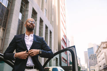 Portrait of a handsome young businessman getting down from car on road and buttoning his jacket. African man standing by his car in city. - JLPSF19791