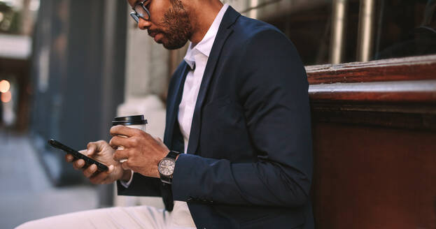 Afrikanischer Geschäftsmann, der draußen mit einem Kaffee sitzt und ein Mobiltelefon benutzt. Ein Mann im Anzug entspannt sich draußen an der Straße, schreibt eine SMS auf seinem Handy und trinkt Kaffee. - JLPSF19794