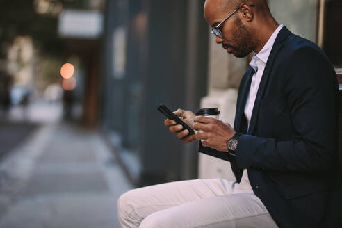 Seitenansicht eines afrikanischen Mannes im Anzug, der an der Straße sitzt, telefoniert und eine Tasse Kaffee in der Hand hält. Geschäftsmann, der sich mit Telefon und Kaffee im Freien entspannt. - JLPSF19795