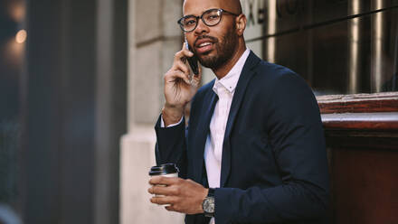 Portrait of handsome young businessman making a phone call while sitting outdoors with a coffee. Man in business suit talking over cell phone. - JLPSF19797