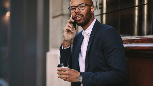 Porträt eines gutaussehenden jungen Geschäftsmannes, der mit einem Kaffee im Freien sitzt und telefoniert. Mann im Geschäftsanzug, der über ein Mobiltelefon spricht. - JLPSF19797