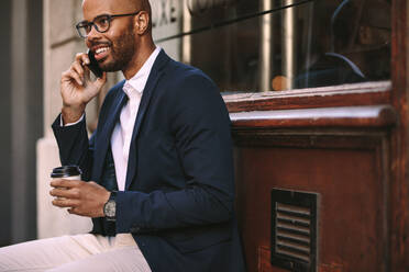 Glücklicher afrikanischer Mann, der sich mit einem Kaffee im Freien entspannt und mit seinem Handy telefoniert. Junger Mann im Business-Anzug sitzt draußen und telefoniert. - JLPSF19798