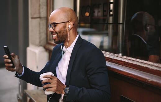 Glücklicher junger afrikanischer Geschäftsmann beim Pendeln mit einem Smartphone. Afrikanischer Geschäftsmann mit Kopfhörern, der im Freien sitzend einen Videoanruf tätigt. - JLPSF19801