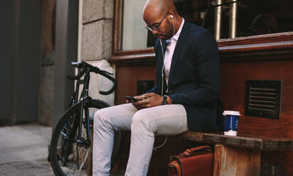 Ein glatzköpfiger afrikanischer Geschäftsmann sitzt im Freien auf einer Bank und telefoniert. Ein Mann im Anzug trägt Kopfhörer und hört Musik auf seinem Smartphone. - JLPSF19802
