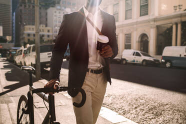 Ausschnitt eines jungen Geschäftsmannes, der mit dem Fahrrad zur Arbeit fährt. Ein Mann im Anzug mit einer Tasse Kaffee in der Hand geht mit seinem Fahrrad durch die Straße. - JLPSF19805