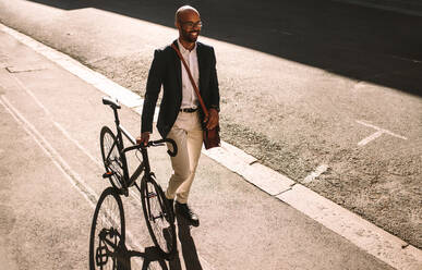 Gut aussehender junger erwachsener Mann im Anzug, der mit seinem Fahrrad im Freien spazieren geht. Afrikanischer Geschäftsmann, der mit seinem Fahrrad ins Büro fährt. - JLPSF19806