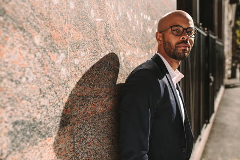 Portrait of handsome young man wearing suit leaning to a wall outdoors and looking at camera. African businessman with beard wearing eyeglasses relaxing outdoors. - JLPSF19808