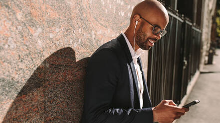 Smiling young businessman leaning on a wall while standing outdoors using mobile phone. Man in suit wearing earphones looking at his smart phone. - JLPSF19809