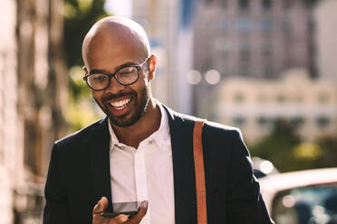 Lächelnder junger afrikanischer Geschäftsmann beim Pendeln mit einem Mobiltelefon, während er im Freien spazieren geht. Glatzköpfiger Mann im Anzug, der in der Stadt spazieren geht und mit einem Mobiltelefon spricht. - JLPSF19812