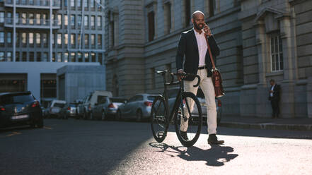 Junger Geschäftsmann, der mit einem Fahrrad im Freien spazieren geht und ein Mobiltelefon benutzt. Afrikanischer Mann, der mit dem Fahrrad zur Arbeit fährt und über ein Mobiltelefon auf einer Stadtstraße spricht. - JLPSF19814