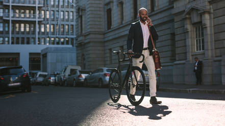 Young businessman walking outdoors with a bicycle and using mobile phone. African man going to work on bike talking over cell phone on city street. - JLPSF19814