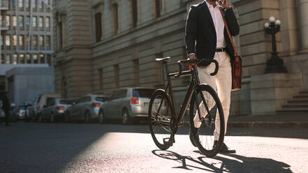 Cropped image of businessman walking on city street with a bicycle and talking on mobile phone. Man in formal wear going to work with cycle using phone. - JLPSF19815