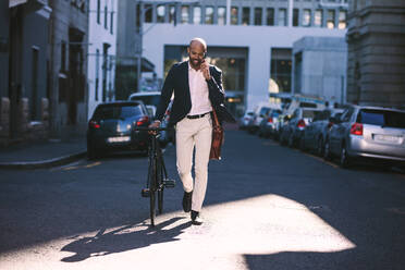 Gut aussehender junger afrikanischer Mann im Anzug, der telefoniert, während er morgens mit dem Fahrrad zur Arbeit fährt. Geschäftsmann, der mit dem Fahrrad zur Arbeit fährt. - JLPSF19816